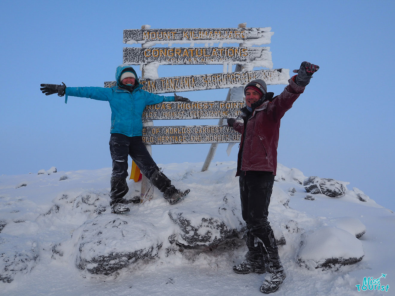 author of the post with husband in winter clothing stand triumphantly with raised arms in front of the Mount Kilimanjaro summit sign, surrounded by snow and ice.