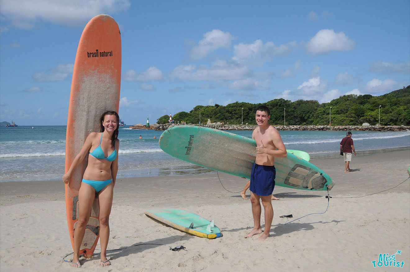 author of the post with husband standing on a sandy beach holding surfboards, with the ocean and other beachgoers in the background.