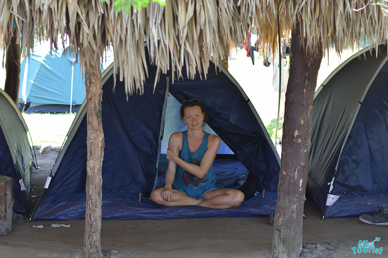 author of the post sits at the entrance of a tent under a thatched roof, surrounded by other tents and trees.