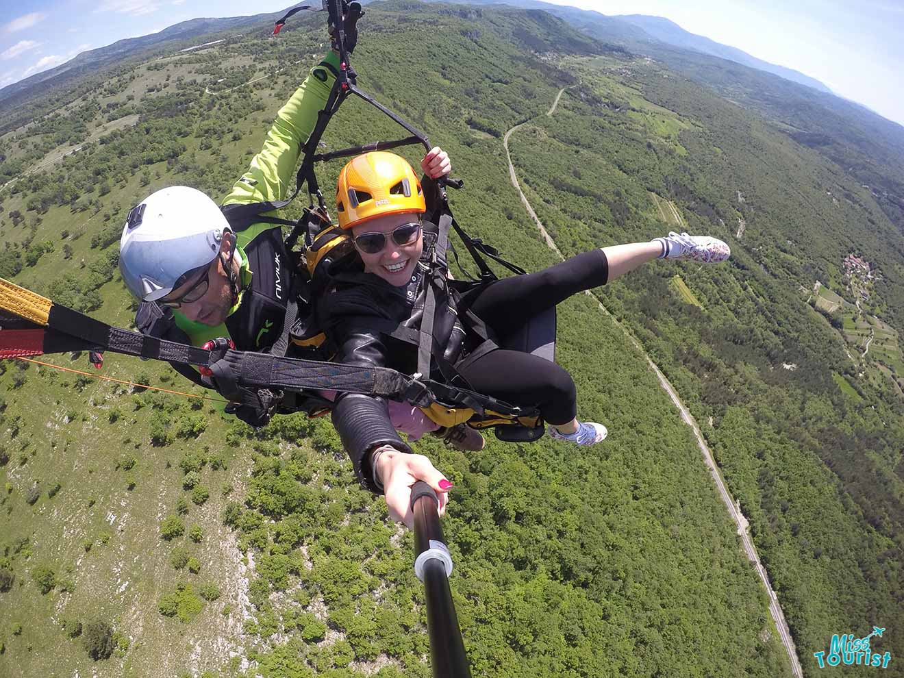 Two individuals paragliding over a lush green landscape with mountains in the background. One is holding a selfie stick taking a photo. Both are wearing helmets.