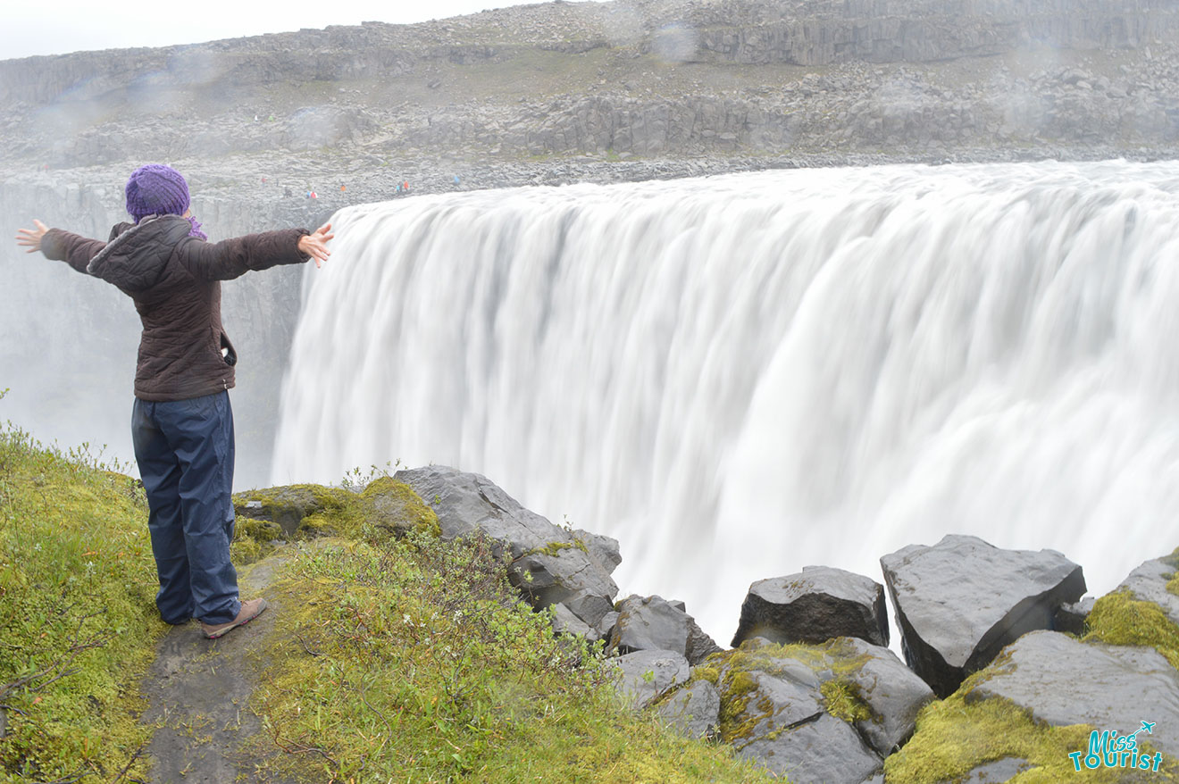 author of the post standing on grassy edge with arms outstretched near powerful, misty waterfall cascading over rocky cliff.