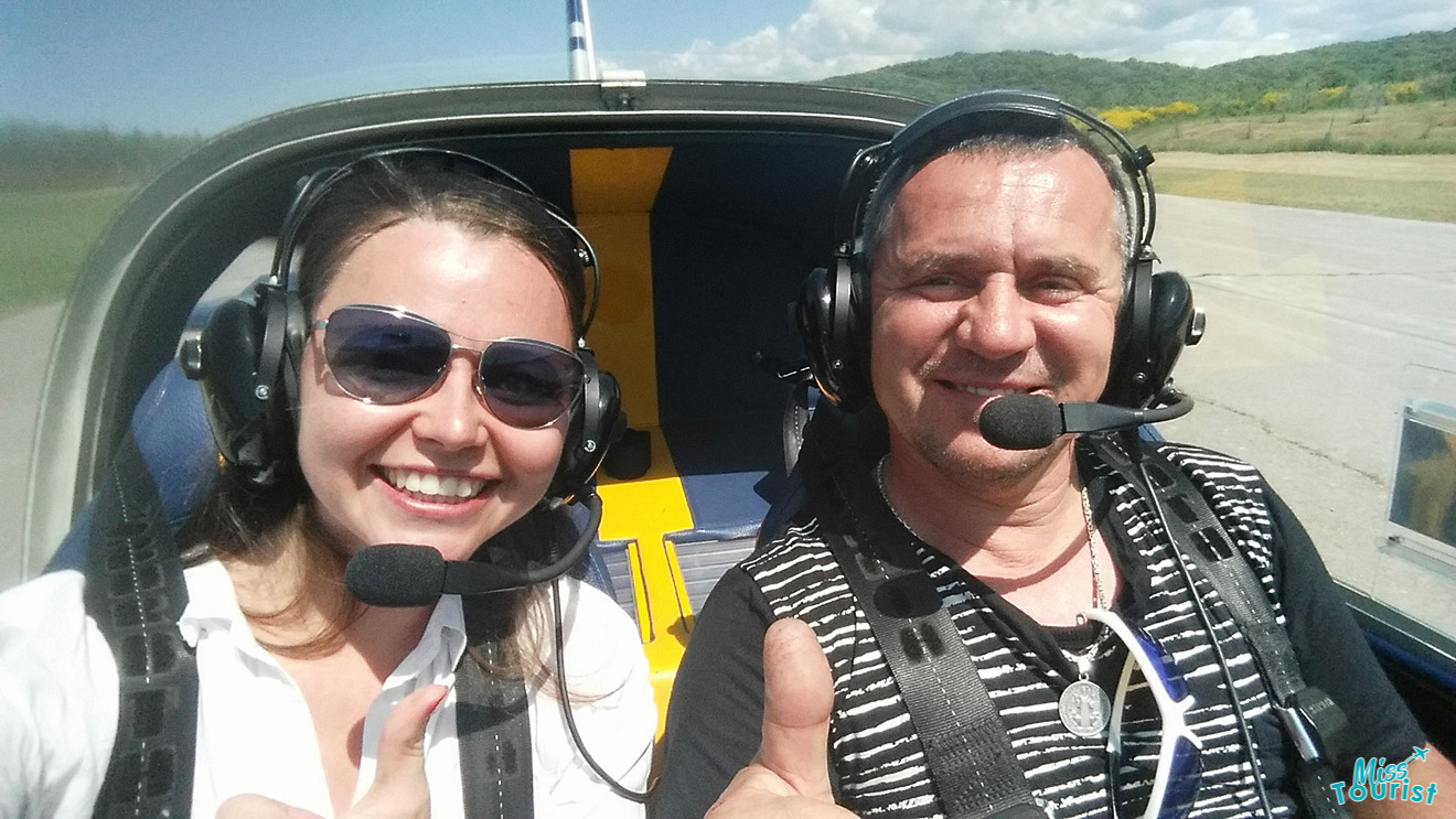 author of the post in sunglasses and pilot wearing headsets are sitting in a small aircraft's cockpit with thumbs up. A rural landscape is visible in the background.