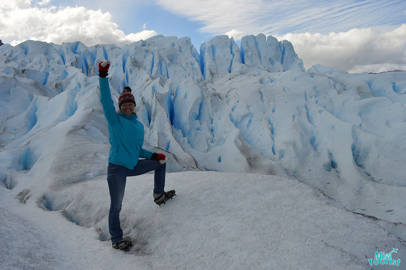 author of the post wearing winter clothing stands with one foot on a snow-covered peak, raising one arm, in front of a large, icy glacier under a cloudy sky.