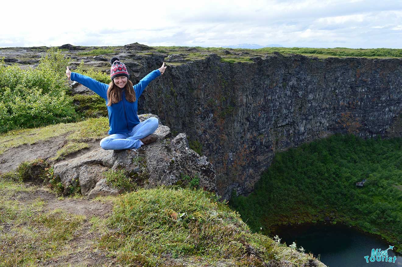 author of the post wearing a blue jacket and purple hat sits cross-legged on a rocky ledge with arms outstretched, overlooking a large canyon with greenery.