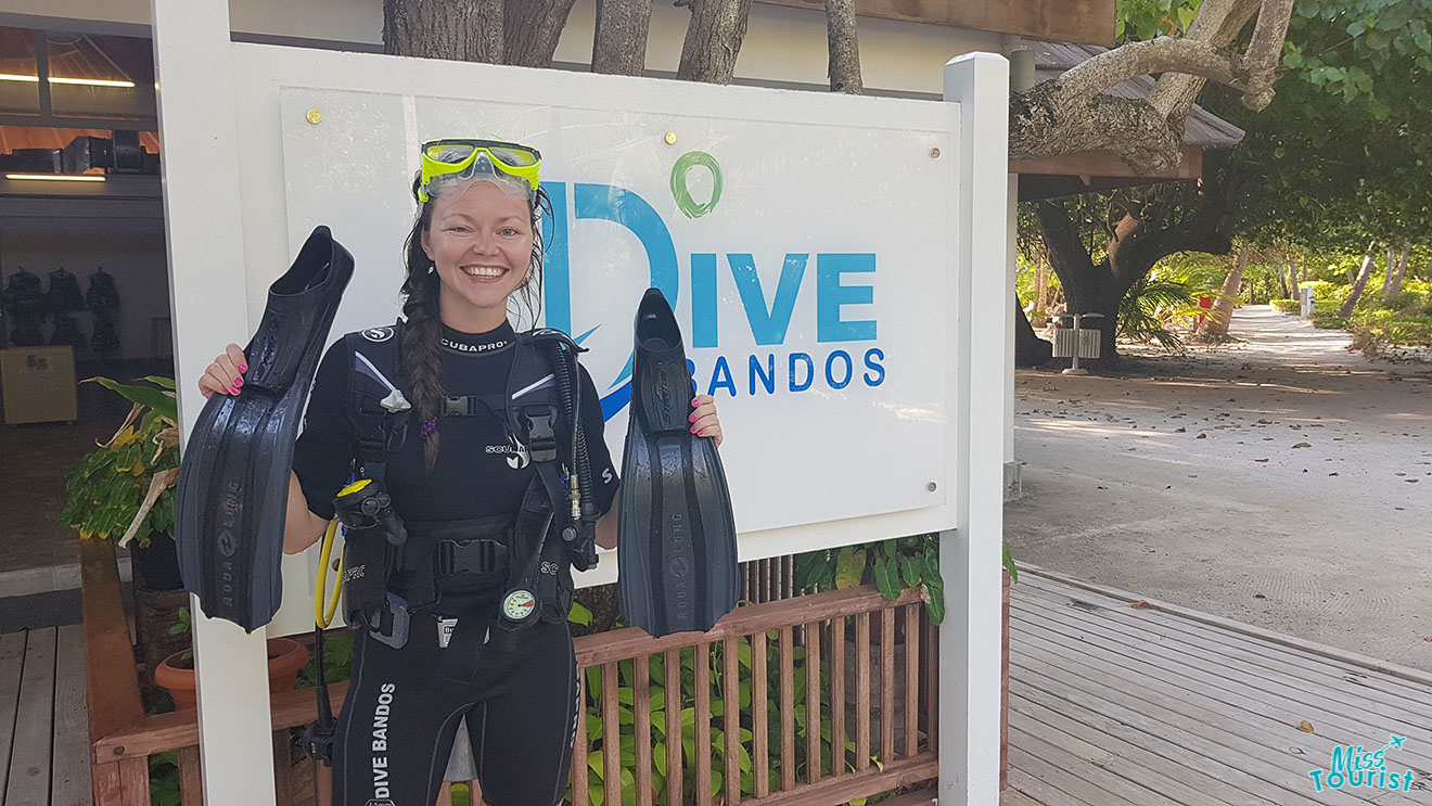 author of the post dressed in scuba diving gear, holding a pair of flippers, stands smiling in front of a Dive Bandos sign.