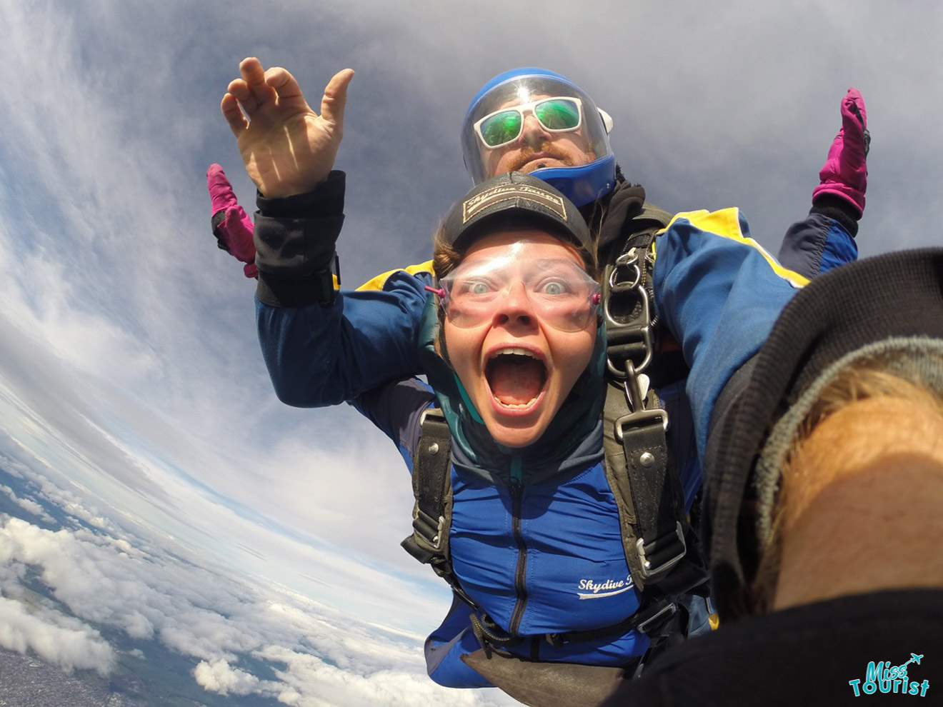 A tandem skydiving pair in mid-air with the instructor behind, author of the post in front wears a blue suit, pink gloves, and goggles, with a wide-open mouth, and clouds visible below.