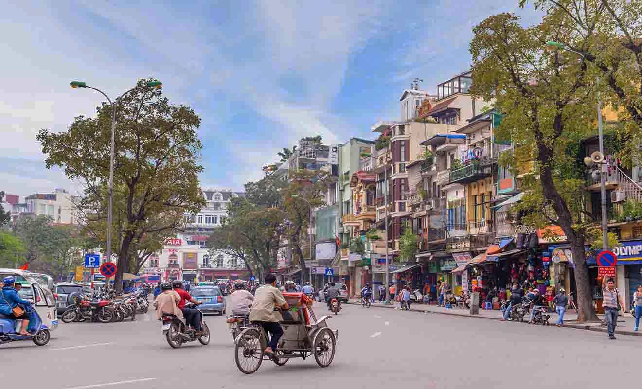 Streets of Hanoi bustling with traffic, cyclists, and pedestrians, lined with shops and autumnal trees, capturing the lively urban spirit