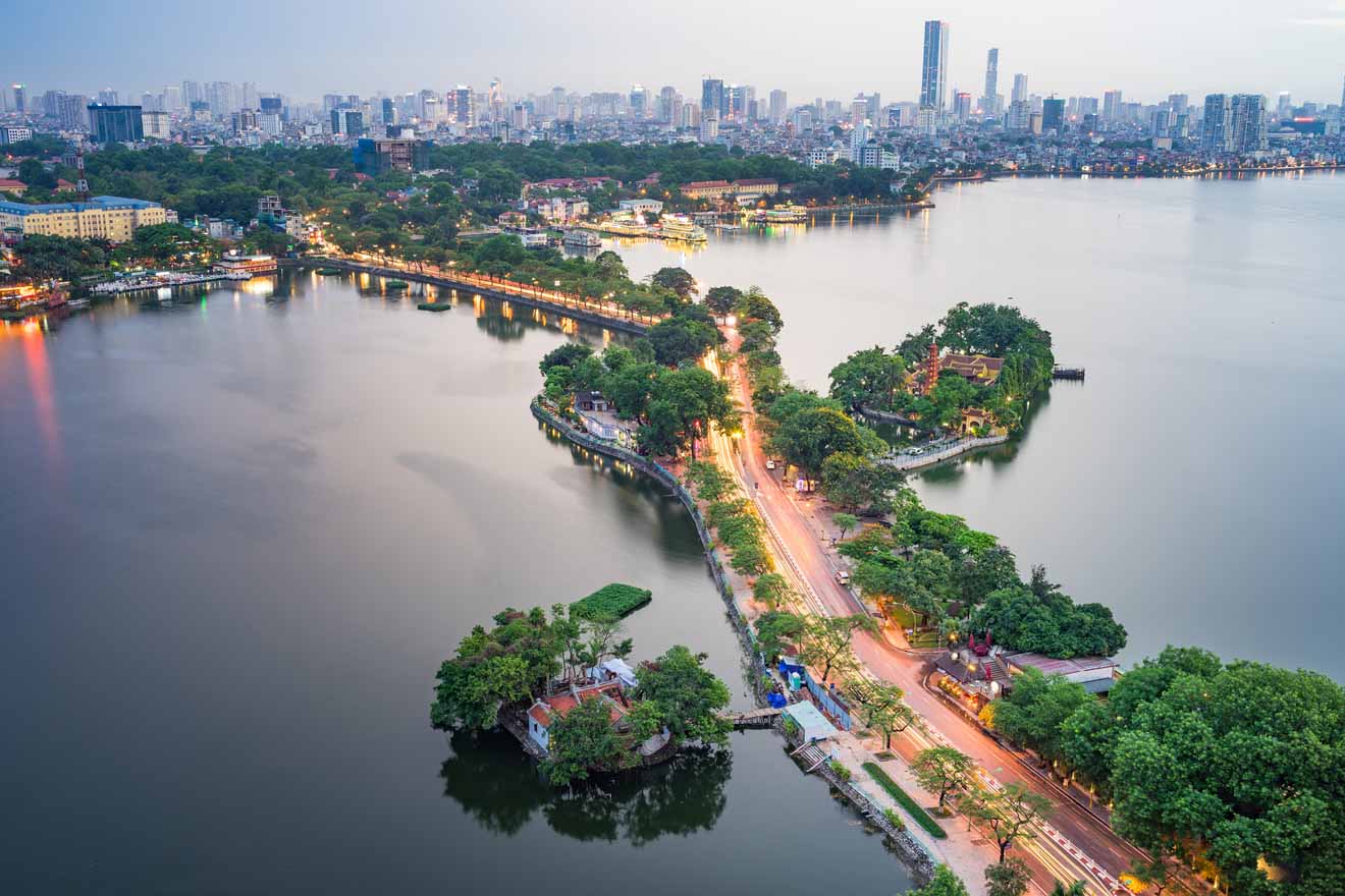 Aerial view of Hanoi with a serene lake at twilight, a well-lit road curving between patches of greenery, reflecting the city's mix of tranquility and urban development