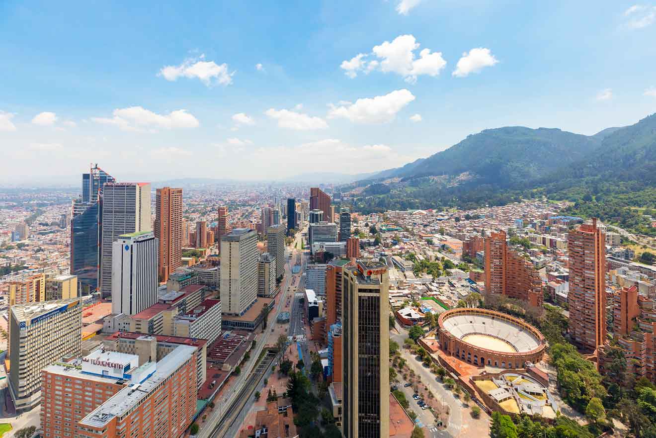 Aerial view of a bustling cityscape with tall buildings, a circular structure resembling a bullring, and a mountainous backdrop under a partly cloudy sky.