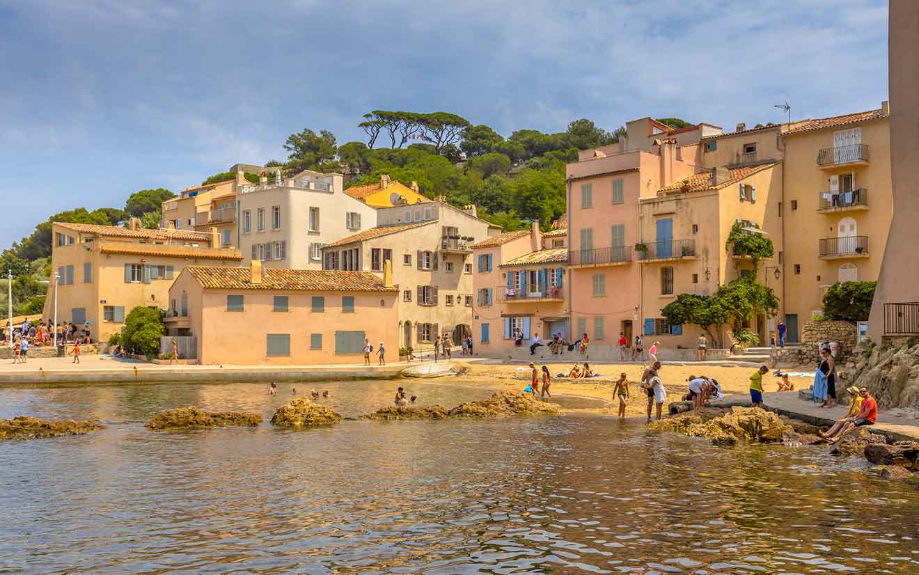 A charming beach scene in Saint-Tropez, with people enjoying the sandy shore near pastel-colored houses and greenery, under a clear sky