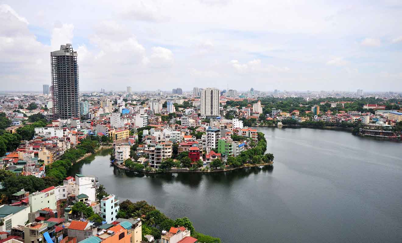 Panoramic view of Hanoi's dense cityscape with buildings clustered around a calm lake, under a cloudy sky, depicting urban life amidst nature