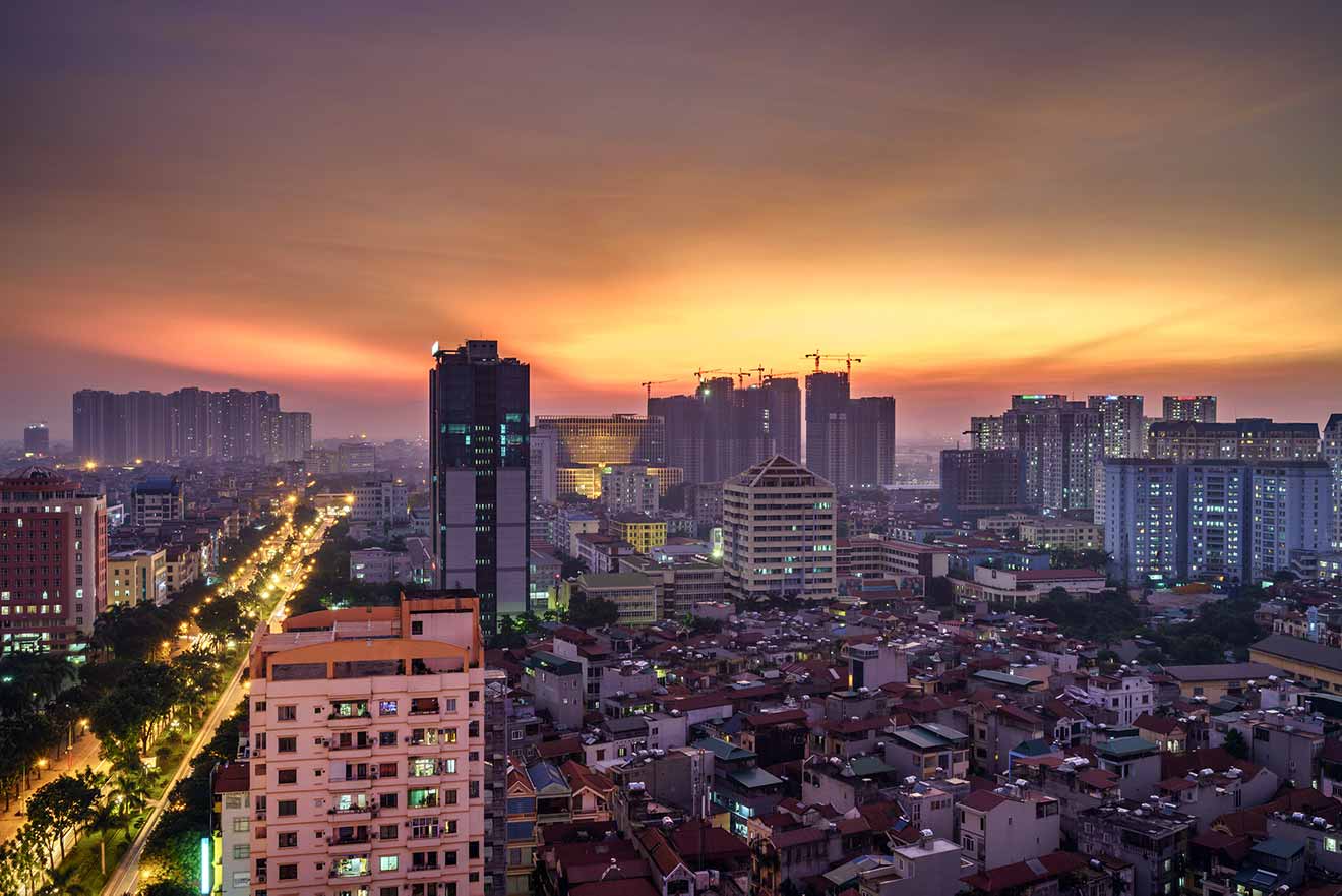 Evening descends on Hanoi, the sky a tapestry of orange hues above the city's illuminated skyline, with busy streets signaling the city's vibrant nightlife
