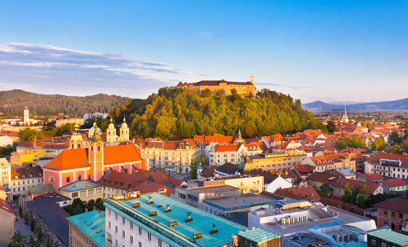 Aerial view of a cityscape featuring a hilltop castle surrounded by lush greenery. Below are colorful buildings with red roofs, including a prominent church with twin towers, all under a clear blue sky.