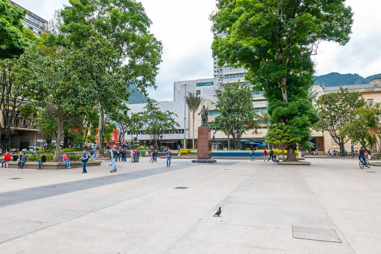 A spacious public square with trees, people walking, and a statue in the center. Buildings and a mountain are visible in the background.