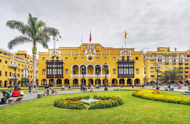 people walking around a big yellow building with a flag and a park in front of it in Lima, Peru