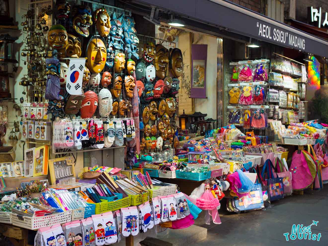 A colorful market stall displaying traditional masks, various souvenirs, stationery, and bags. The shop is named "Aeq Sagig Madang" and has "Miss Tourist" signage.