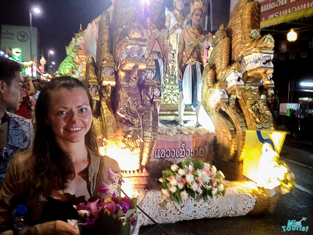 The writer of the post, Yulia Saf, holding flowers stands in front of a richly decorated float featuring traditional Thai figures, participating in a night parade, with the ambient glow