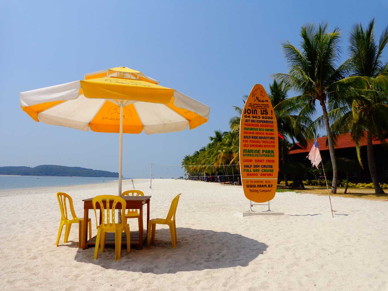 A sunlit beach scene in Langkawi with a large orange and white umbrella, wooden chairs, and a signpost advertising various marine activities