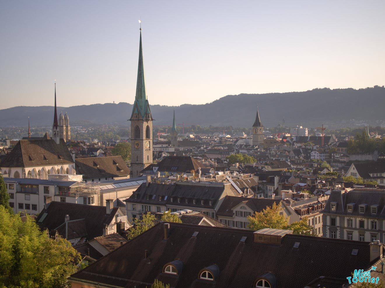 A panoramic view of Zurich's skyline showing the Fraumünster and St. Peter's churches with their iconic clock towers, set against a backdrop of distant hills, under a clear sky at dusk