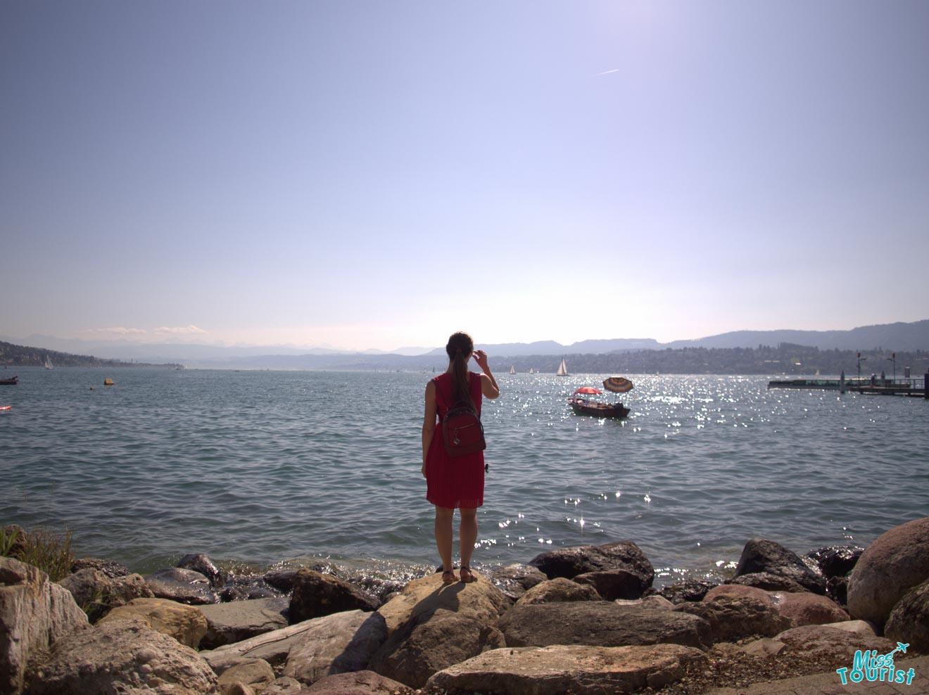 A lone individual with a red backpack stands on the rocky shore of Lake Zurich, gazing out at the shimmering water and distant sailing boats under a hazy sky.