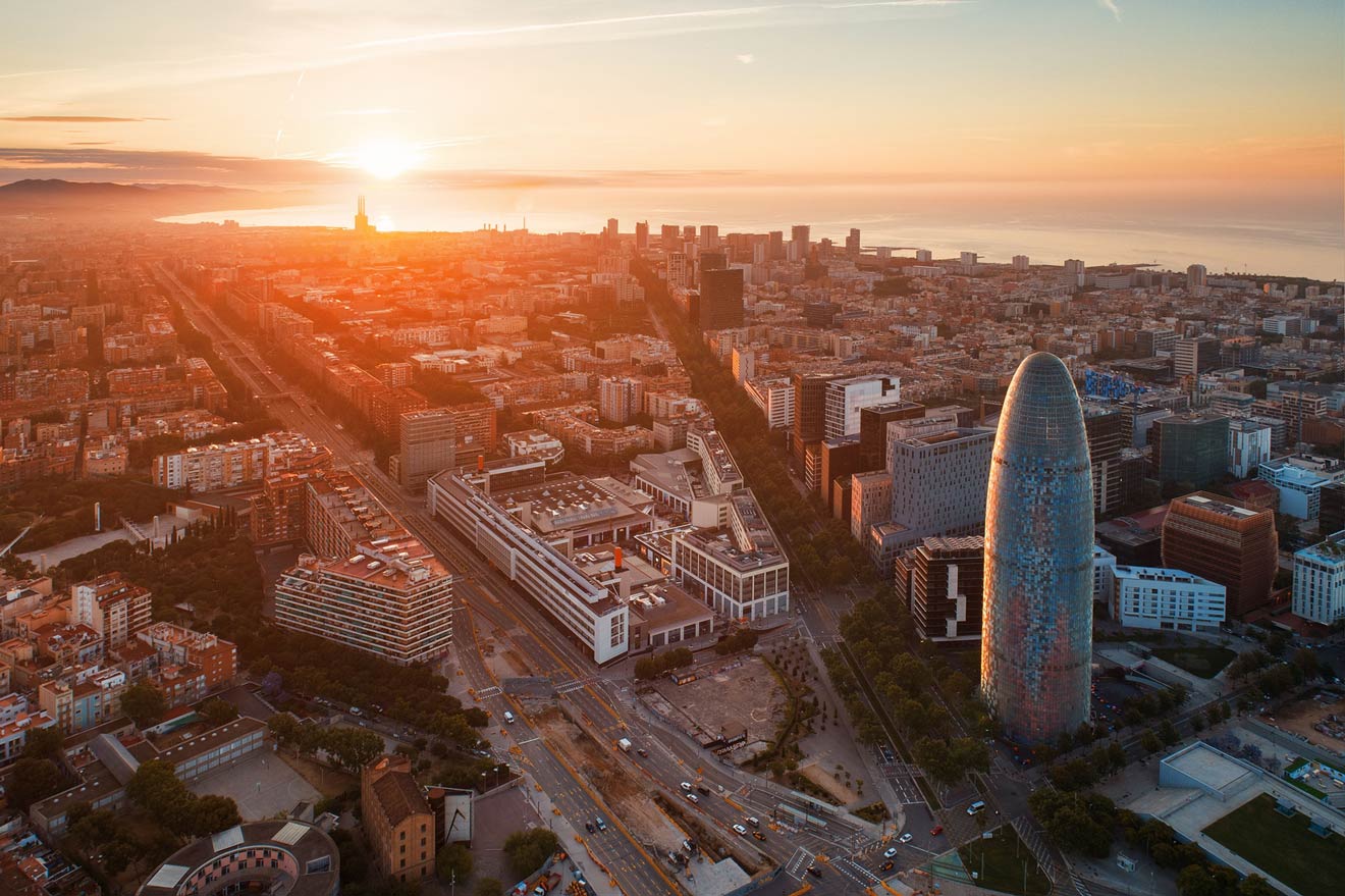 Aerial view of a cityscape at sunset, featuring modern buildings, a distinctive tower, roads, and the coastline in the background.