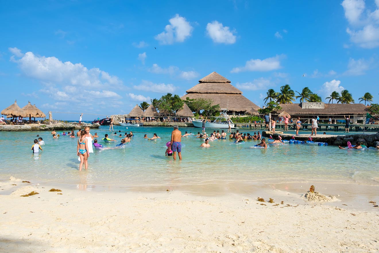 A crowded beach scene with people swimming and lounging in the water, with thatched roof structures and palm trees in the background on a sunny day.