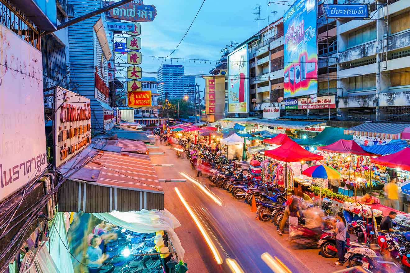 A bustling evening street market in Chiang Mai, Thailand, with vendors under colorful umbrellas, vibrant neon signs in Thai script, and a blur of motion from passing scooters