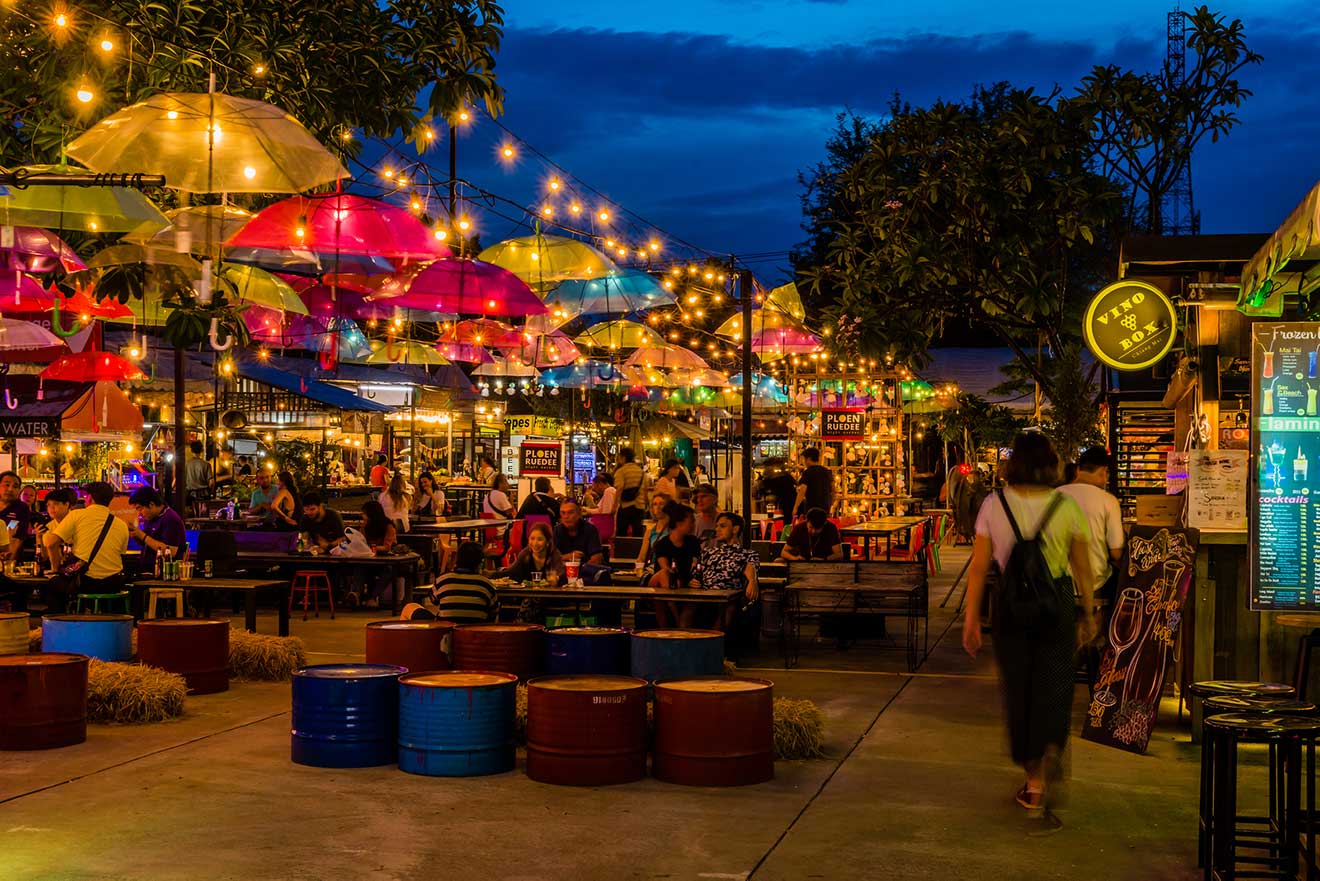 An inviting outdoor night market scene in Chiang Mai, with a canopy of colorful umbrellas and string lights, communal seating on repurposed colorful barrels, and various food stalls