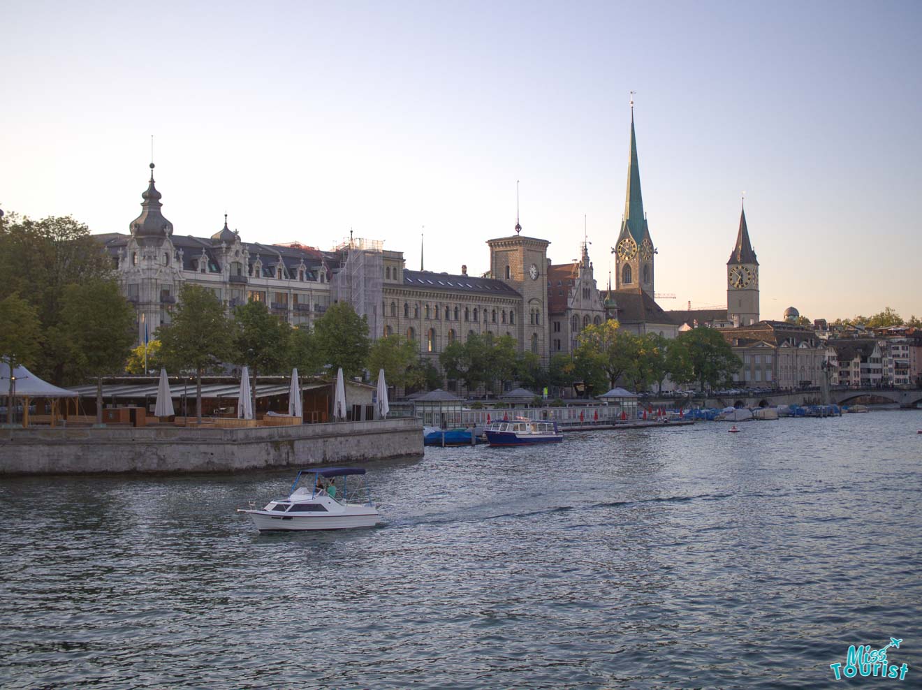 The serene waters of Lake Zurich in the evening, with the spires of Grossmünster church prominent in the background, and boats docked along a tree-lined quay.