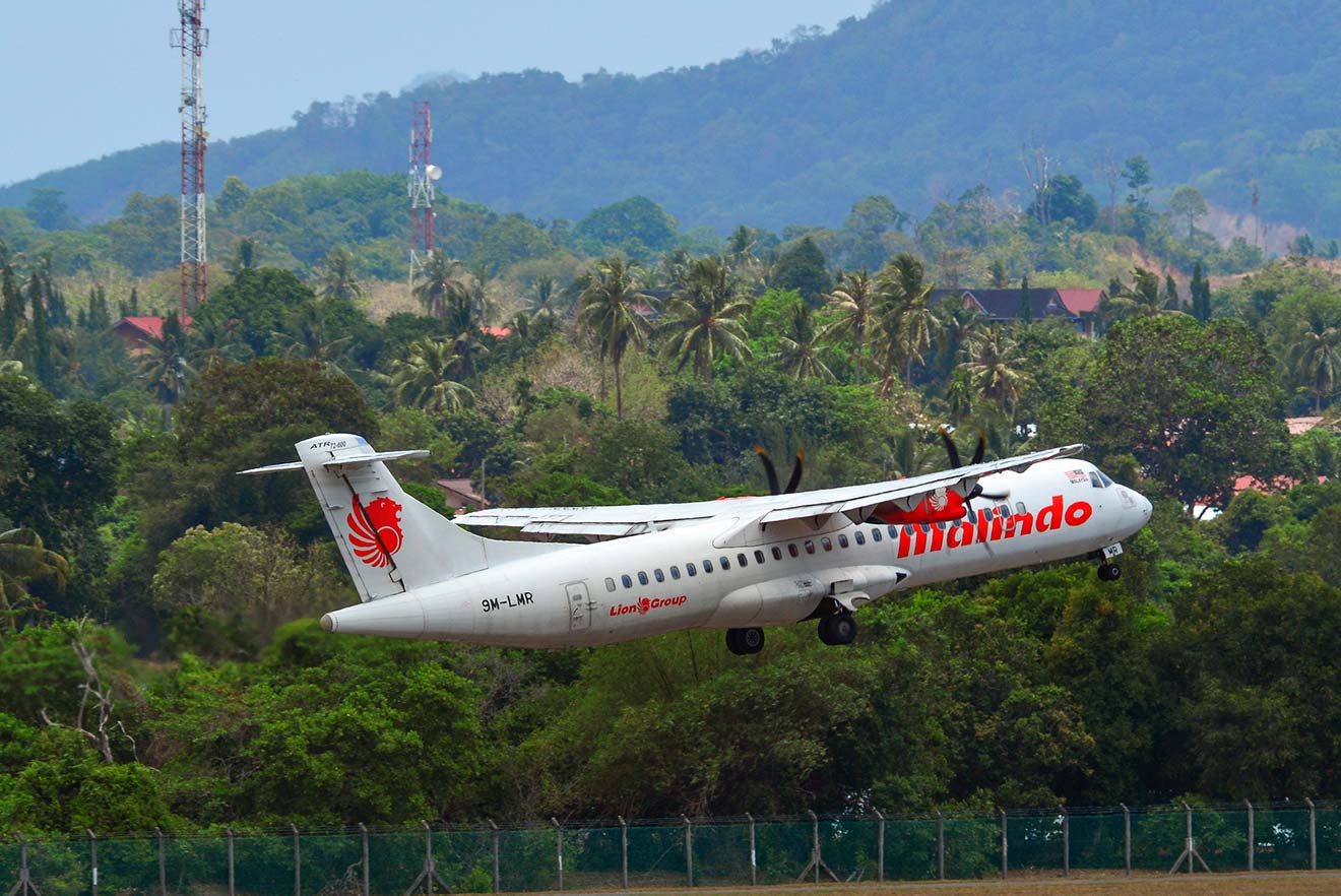 A Malindo Air aircraft in mid-takeoff against a backdrop of tropical greenery and hilly terrain.