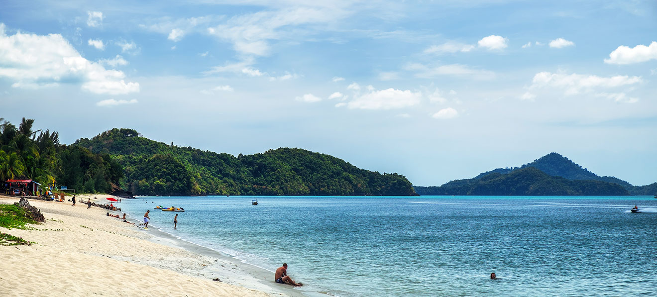 Panoramic view of a sandy beach in Langkawi with visitors, palm trees, and a clear view of a hilly island in the distance under a blue sky with puffy clouds