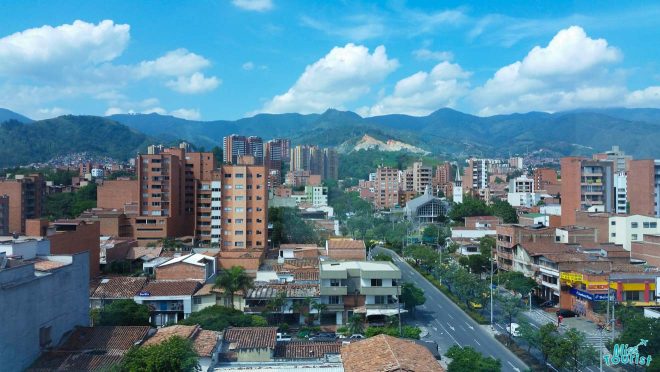 Elevated view of Medellín, Colombia, with a mix of residential and commercial buildings set against a backdrop of green hills under a blue sky with scattered clouds. 