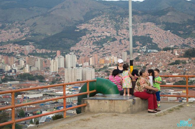 Visitors enjoying the panoramic view of Medellín, Colombia, from a high vantage point. The city's dense urban landscape with numerous buildings stretches into the hills in the background