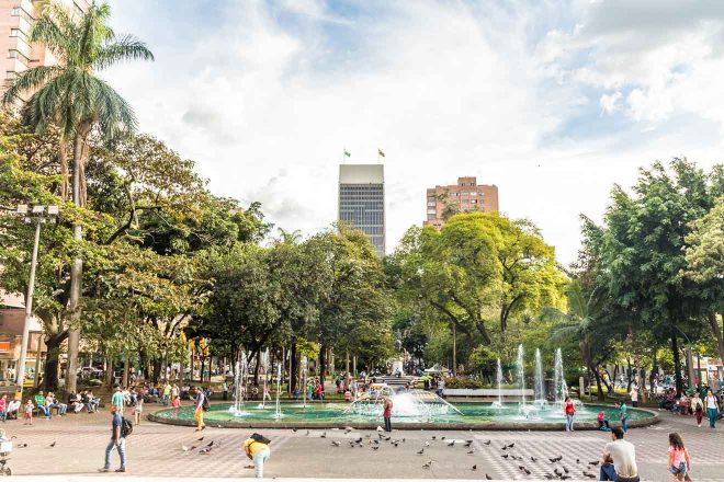 Lush urban park on Plaza Bolivar in Medellin with a central fountain, surrounded by dense foliage and people enjoying the open space, with city skyscrapers in the background
