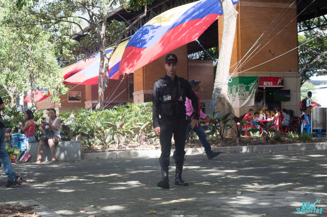 A security guard in uniform stands in a park with people sitting on benches and a large flag in the background, under the shade of trees.