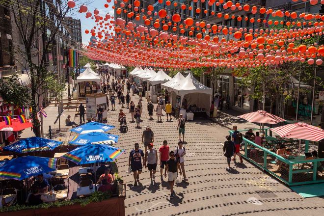 A pedestrian street in Montreal adorned with a canopy of red umbrellas, bustling with market stalls and visitors under a sunny sky.