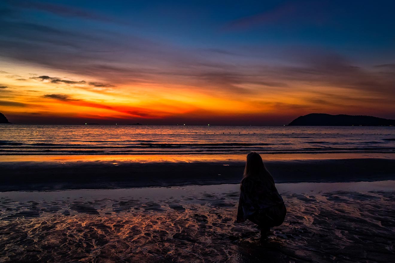 Silhouette of a person sitting on a beach in Langkawi at sunset, with vibrant orange and purple hues reflecting off the water's surface