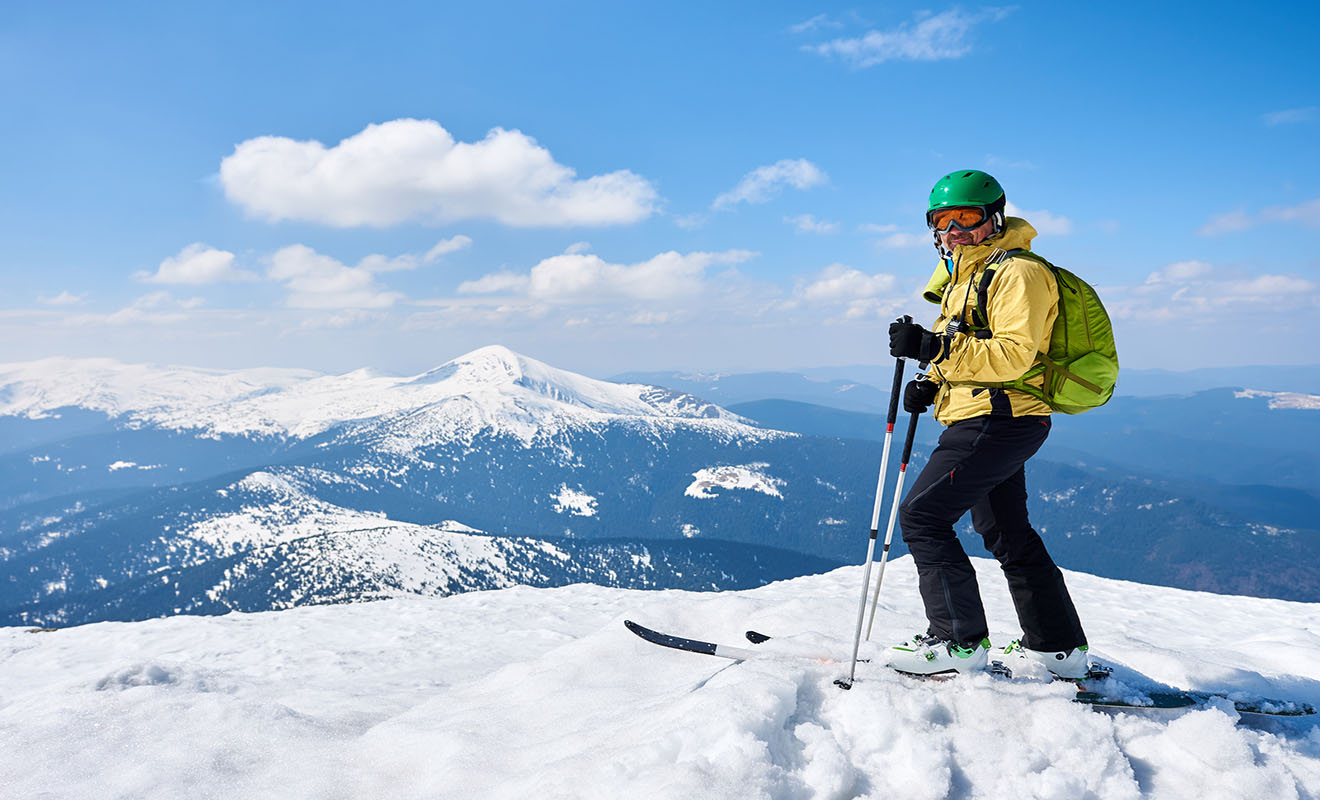 A skier in a yellow jacket and green helmet stands on a snowy mountain peak with snow-covered mountains and a blue sky in the background.