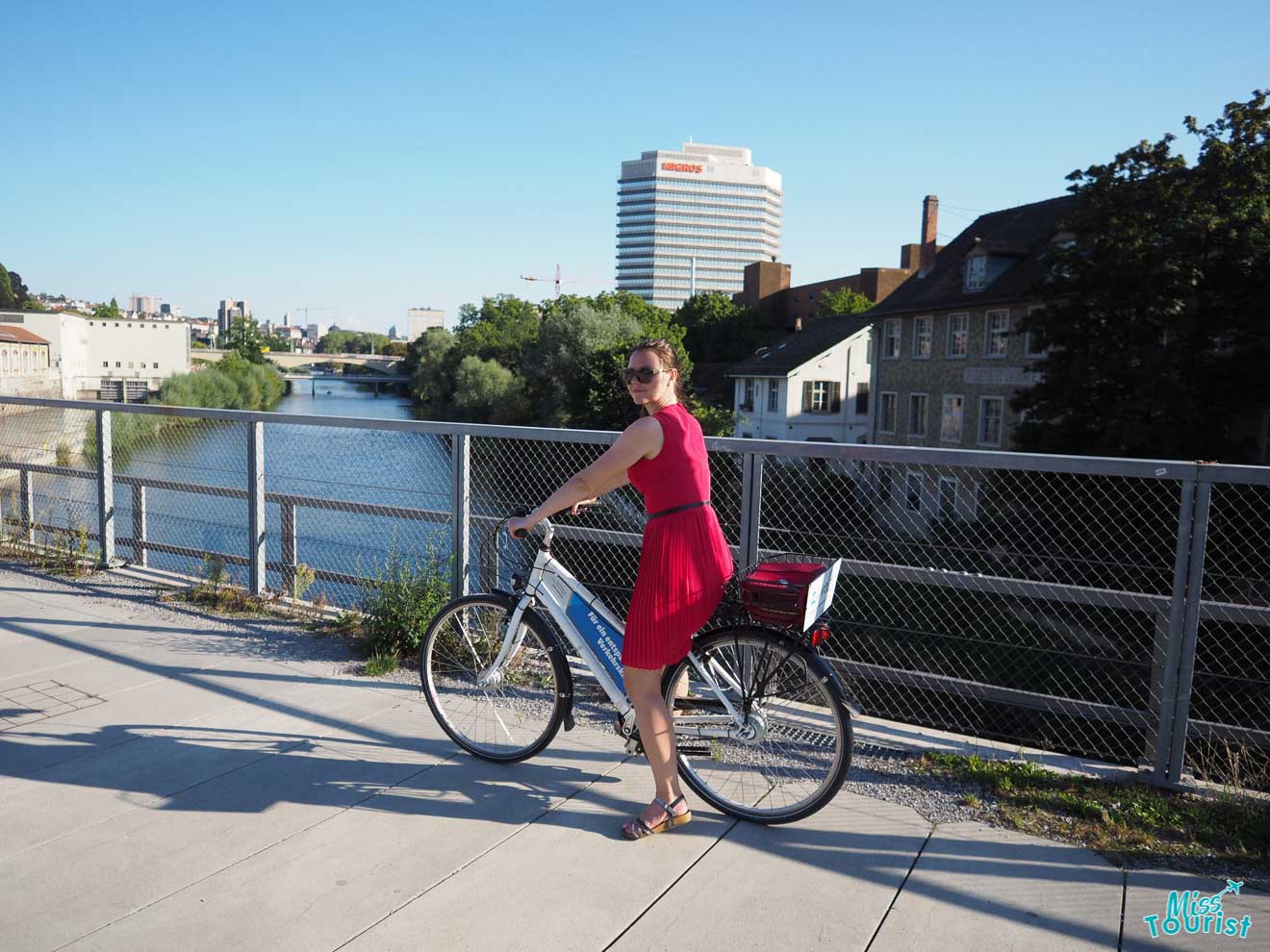 Yulia, the founder of this website, in a red dress stands with a bicycle on a riverside path, with buildings and a river visible in the background.