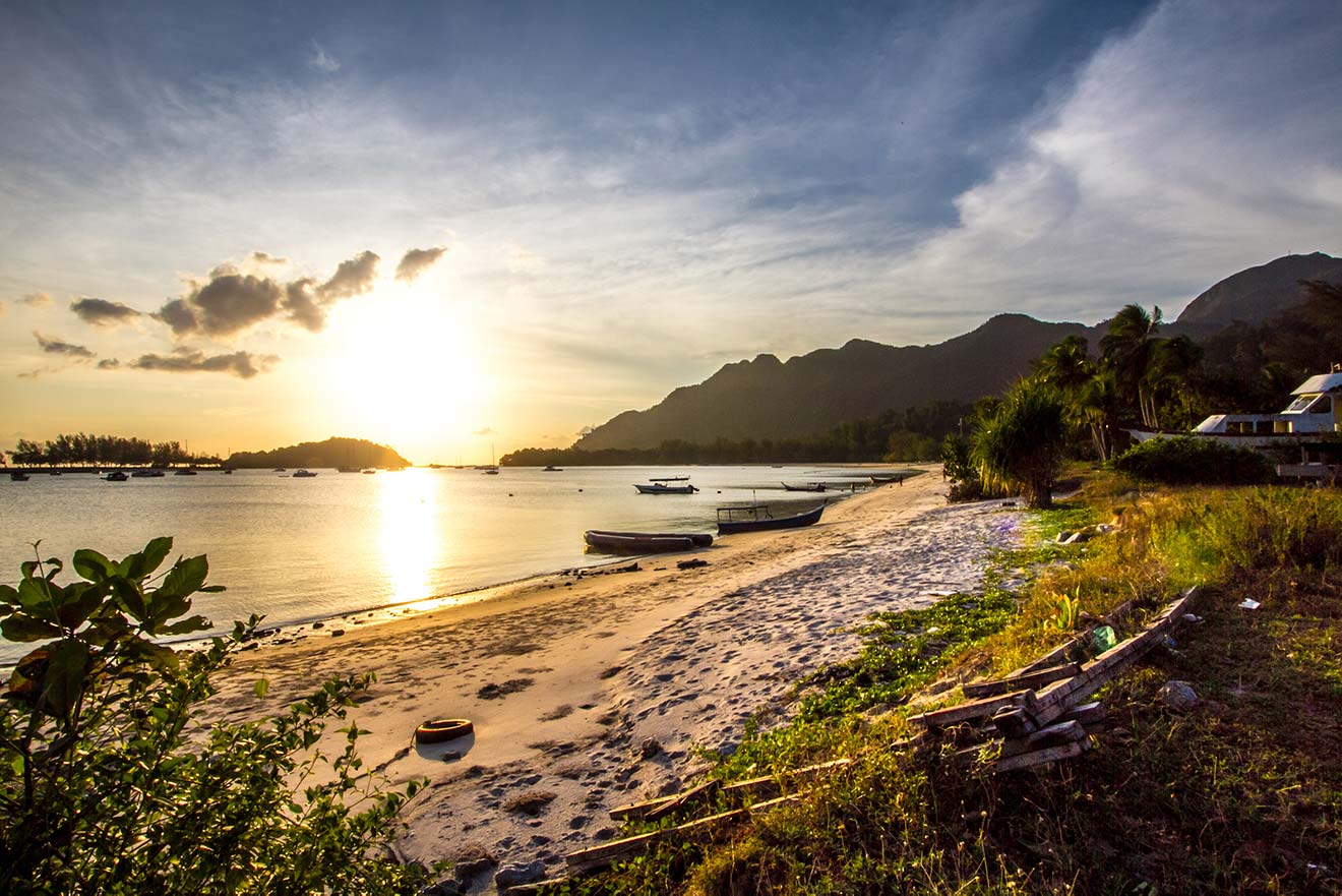 A serene beachscape in Langkawi at sunrise with boats moored near the shore, a mountain backdrop, and a village in the distance.