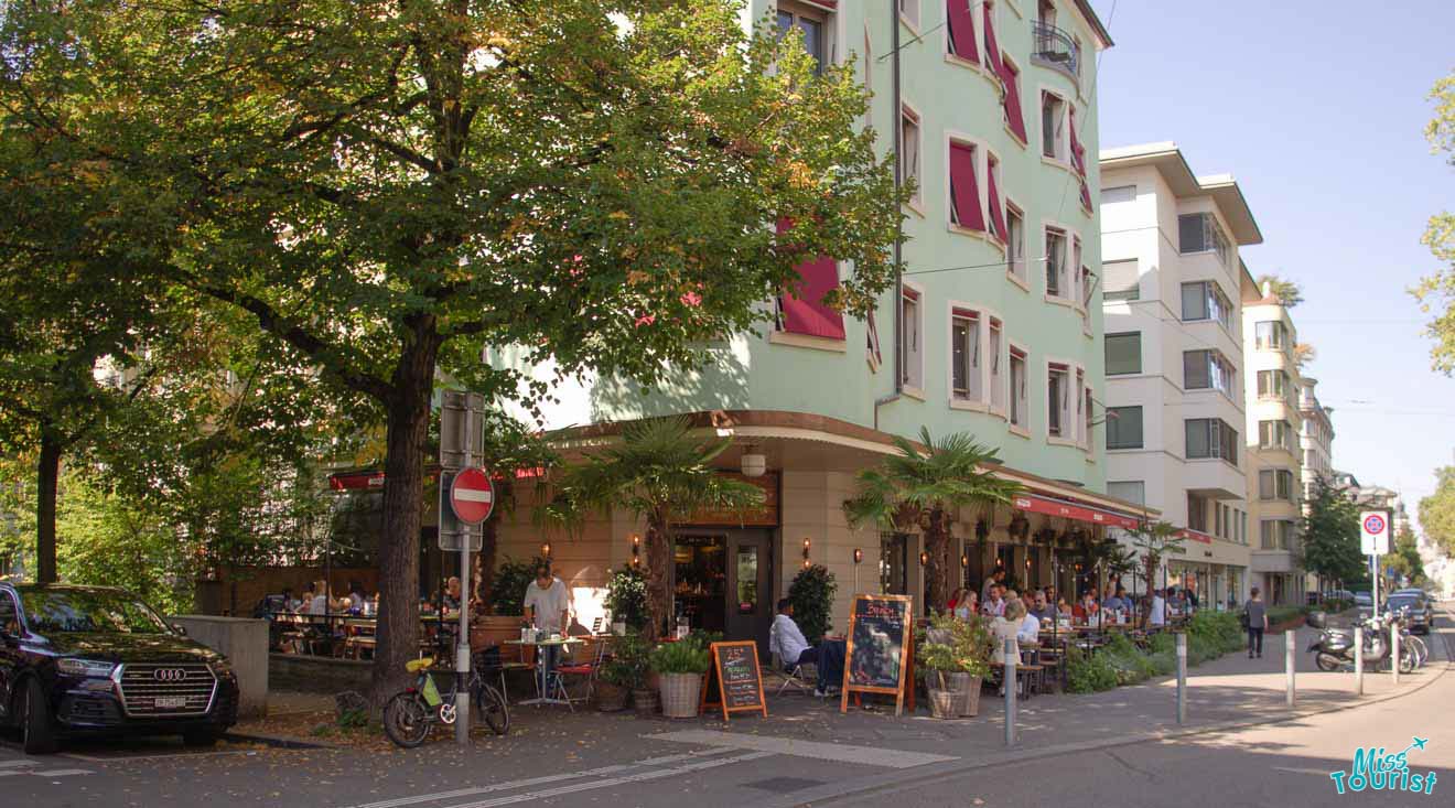 A lively street cafe scene in Zurich with outdoor diners, adjacent to a tree-lined sidewalk and residential buildings on a sunny day.