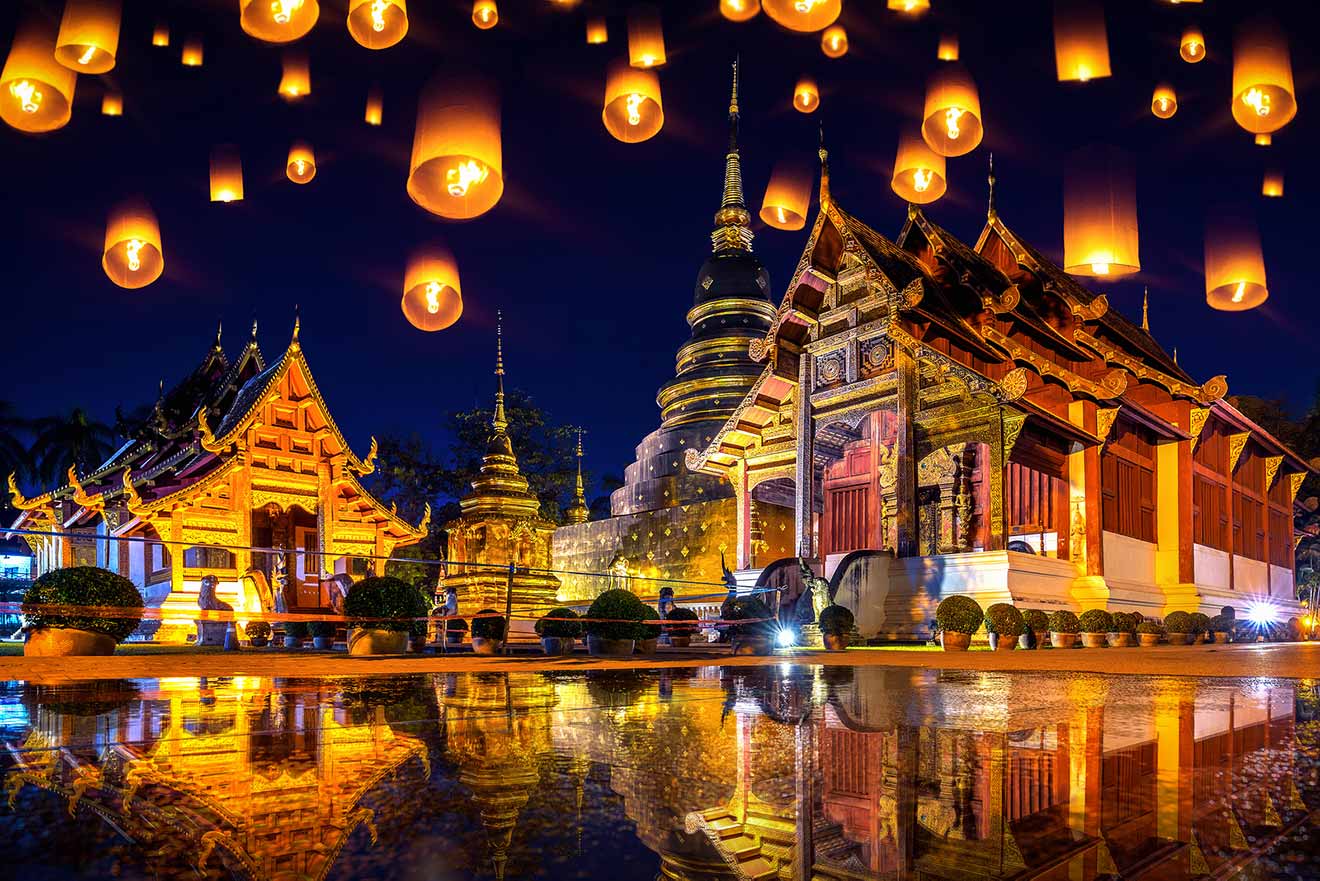 Floating lanterns illuminate a traditional temple complex at night, reflecting in the water below.