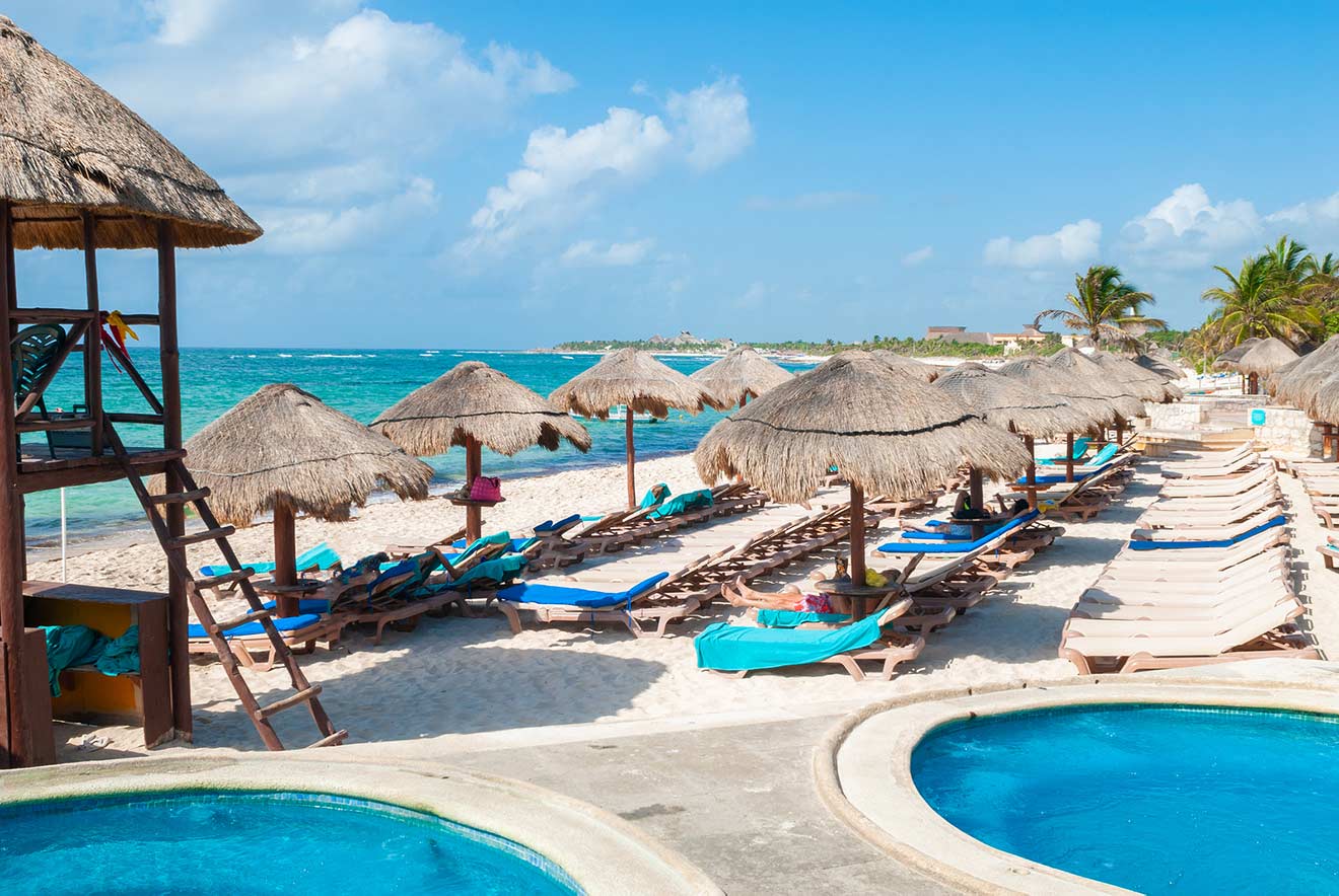 Beach with straw umbrella sunshades, lounge chairs on white sand, lifeguard tower to the left, and two small circular pools in the foreground. Clear blue sea and sky in the background.