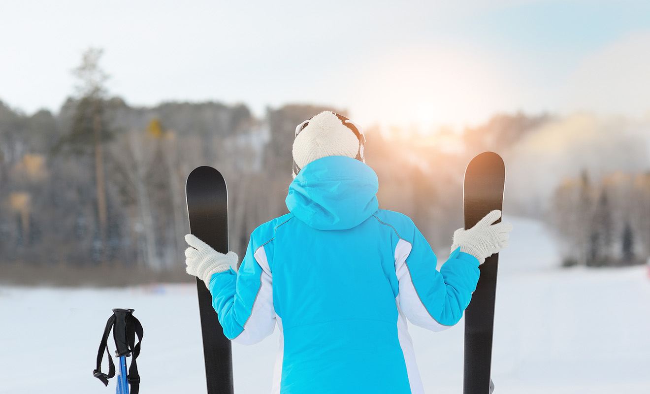 A person in a blue jacket and white hat holds skis, standing on a snowy slope, facing the sunrise. A pair of ski poles is nearby.