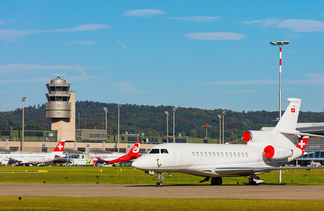Zurich Airport with a variety of planes on the tarmac, including Swiss International Air Lines jets, under a blue sky with the air traffic control tower visible in the background.