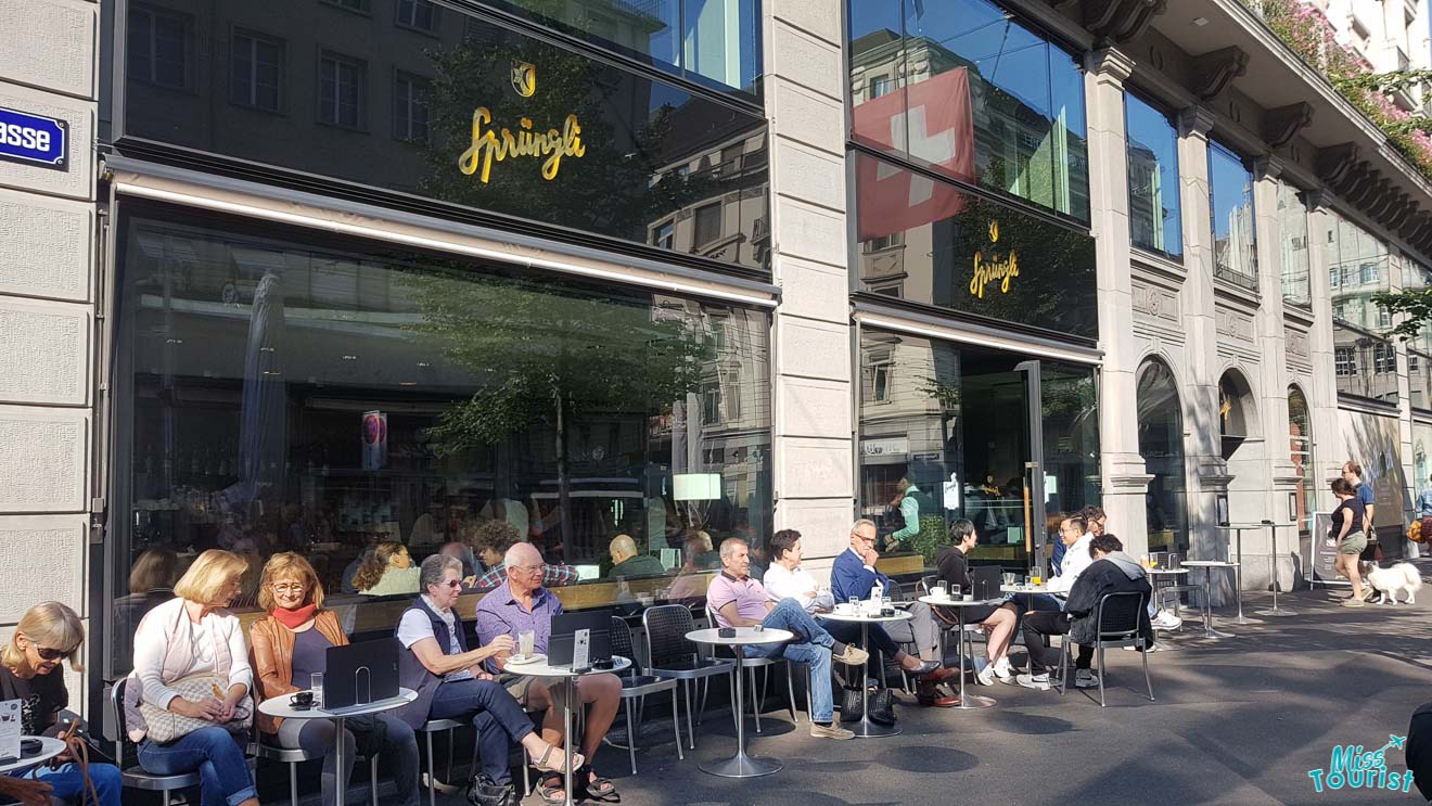 Outdoor seating at the bustling Sprüngli cafe on a sunny day in Zurich, with patrons enjoying coffee and conversation.