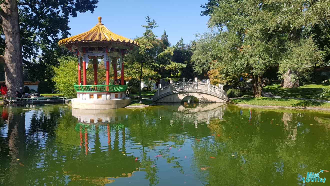 A picturesque Chinese garden with a vibrant red and green pagoda reflected in a tranquil pond, surrounded by lush trees and a stone bridge in Zurich.