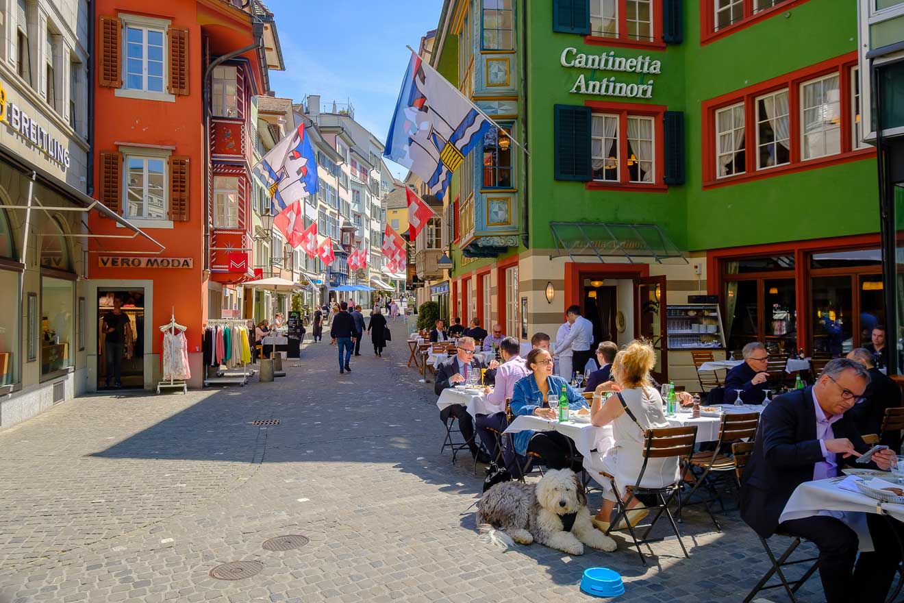 People dining at outdoor tables on a narrow European street surrounded by colorful buildings and flags. A dog sits on the cobblestone pavement near the diners.