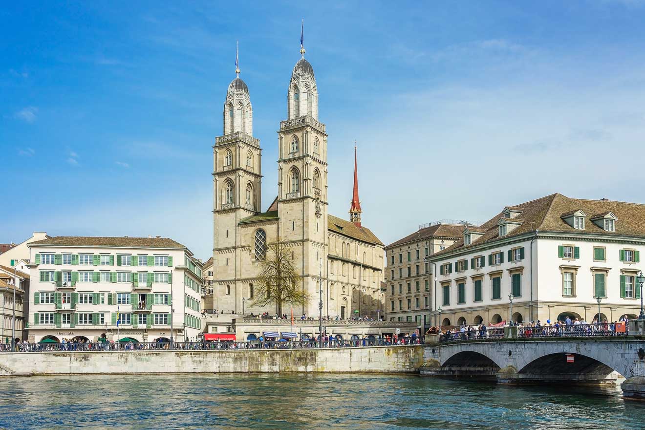 A large historic cathedral with twin towers stands by a river, surrounded by traditional buildings and a stone bridge under a clear blue sky.