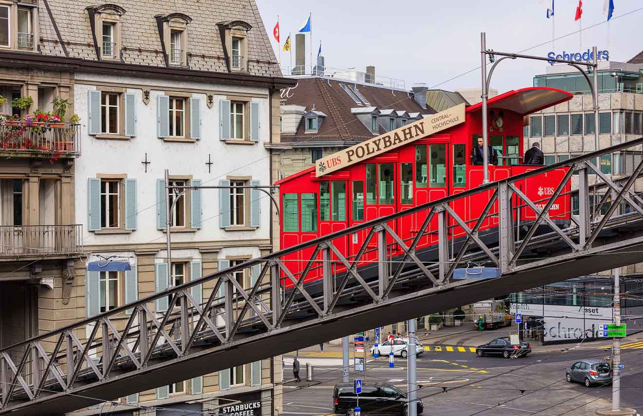 A red funicular labeled "Polybahn" ascends a steep track in an urban area, with buildings and a Starbucks cafe visible in the background.